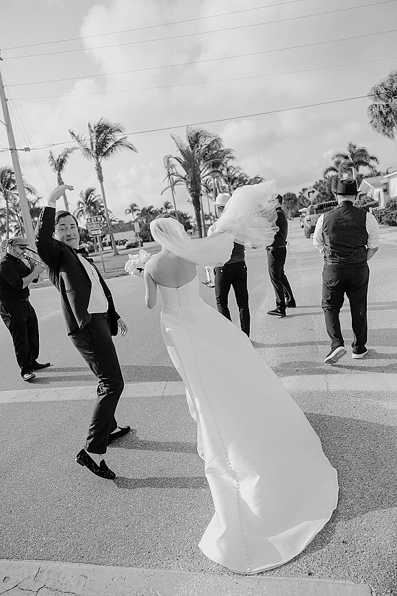 Wedding party portrait in a black and white wedding photo, bride with long train and bouquet walking with groom and groomsmen at a palm-lined intersection