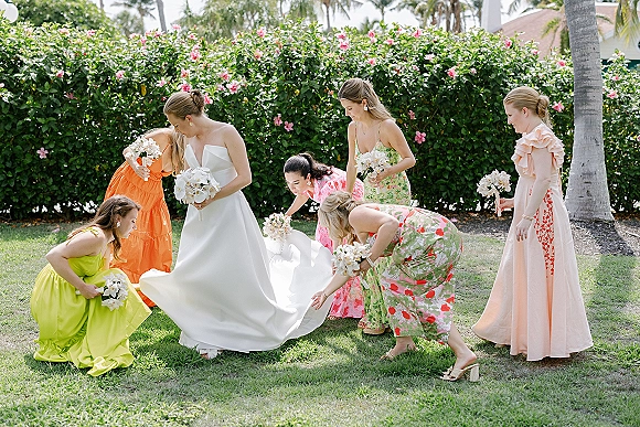 Bridal party photo of bride with bridesmaids adjusting her satin wedding dress train on a grass lawn with palm trees and bouquets