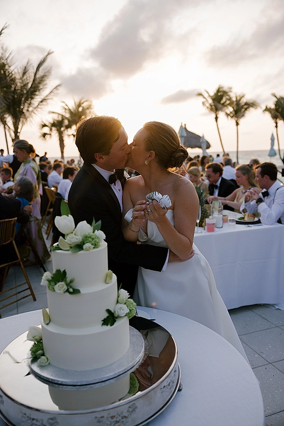 Wedding cake cutting as bride and groom kiss beside a three tier white cake with rose and greenery accents at sunset ocean patio reception
