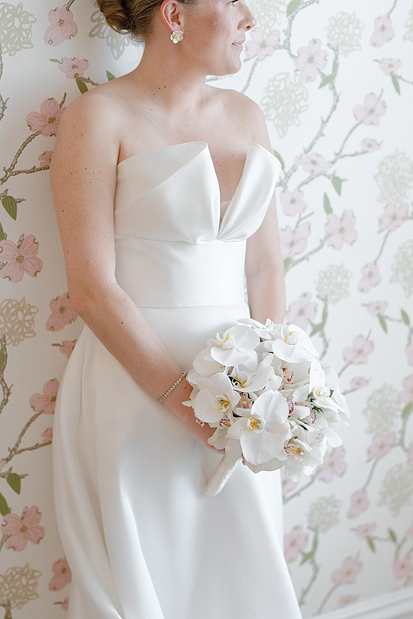 Bridal portrait of a bride in a strapless wedding dress holding a white orchid bouquet, wearing earrings, against floral wallpaper indoors