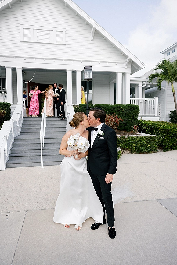 Wedding kiss portrait of bride and groom kissing, her veil and white orchid bouquet against white house porch steps and hedges outdoors