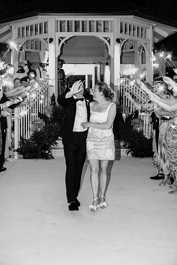 Wedding send off as newlyweds walk through sparklers, bride in mini dress and groom in tuxedo by a gazebo staircase at night