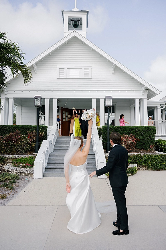 Wedding exit as newlyweds leave ceremony down white chapel steps, bride in veil lifting bouquet beside groom in tux under lanterns