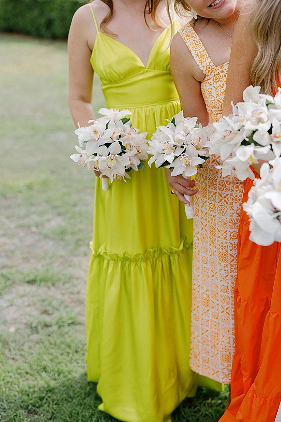 Bridesmaid bouquets of white orchid bridesmaid bouquet stems with ribbon wrap, held by bridesmaids in colorful dresses on a green lawn