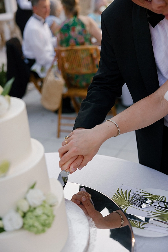 Wedding cake cutting as bride and groom hands hold a knife by a tiered white cake with rose and hydrangea accents on a mirrored stand outdoors