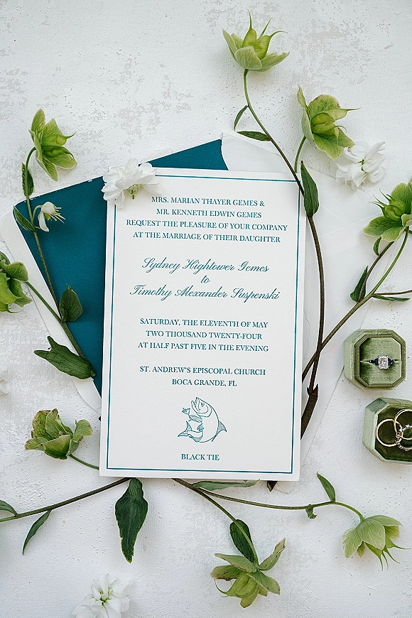 Wedding invitation suite with formal wedding invitations, envelopes, white flowers and greenery, rings and boxes on a white textured surface