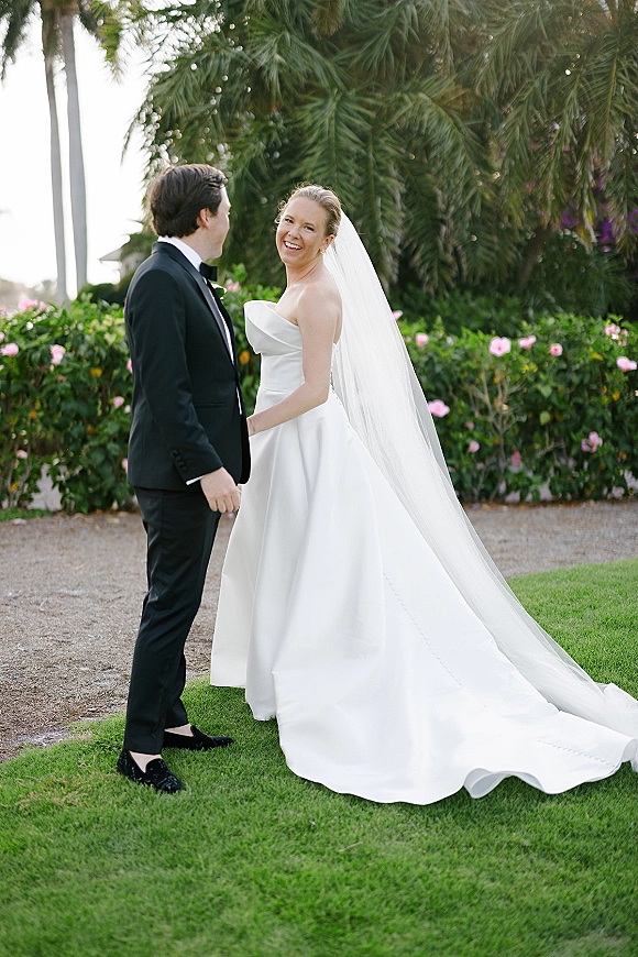 Couple portrait of bride in a strapless dress and long veil holding hands with tuxedoed groom on a lawn with palms and pink flowers