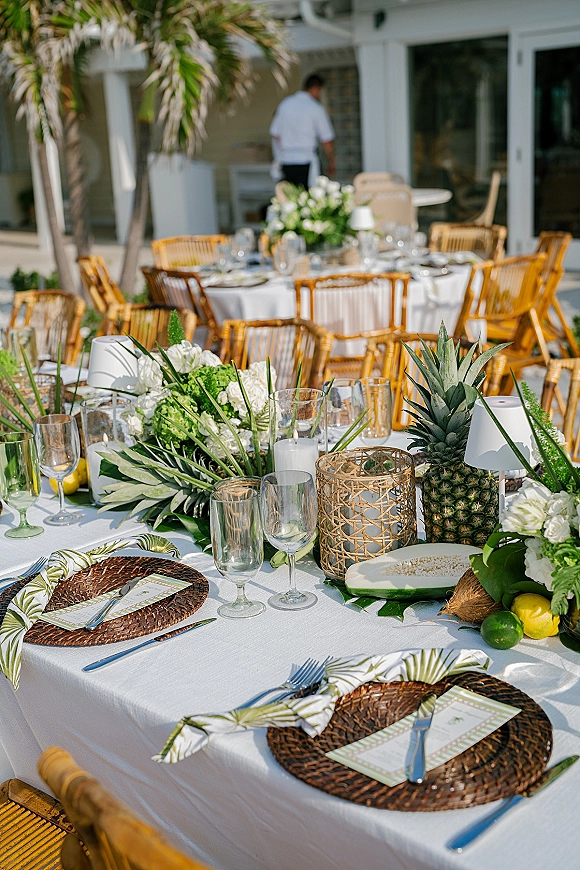 Reception tablescape with tropical wedding tablescape details, rattan charger plates, palm leaf napkins, fruit centerpiece and candles on an outdoor patio