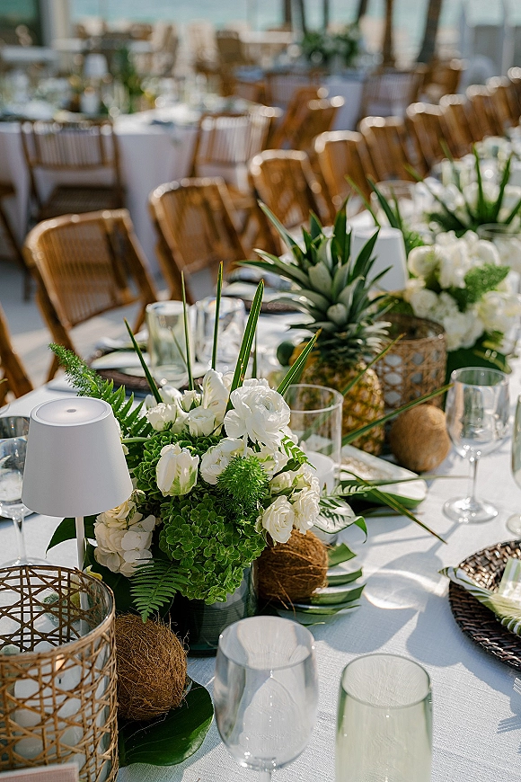 Reception tablescape with tropical wedding tablescape details, green and white florals, palm leaves, pineapples, coconuts, and wicker chargers outdoors