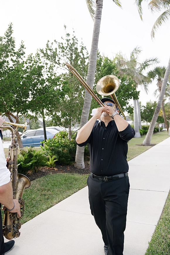 Wedding band performance with a trombone player in black shirt and sunglasses walking past palm trees on a sunny sidewalk outdoors