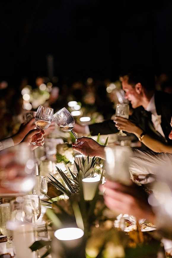 Wedding reception toast as guests cheer and clink wine glasses over a candlelit banquet table with floral greenery centerpiece in dim light