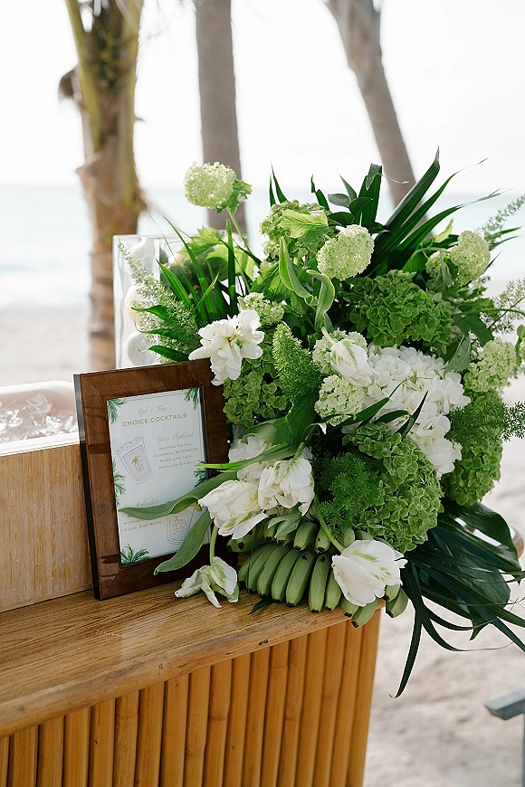 Cocktail bar decor with framed wedding cocktail menu sign, lush tropical greenery and bananas on a wood bar by ocean and palm trees
