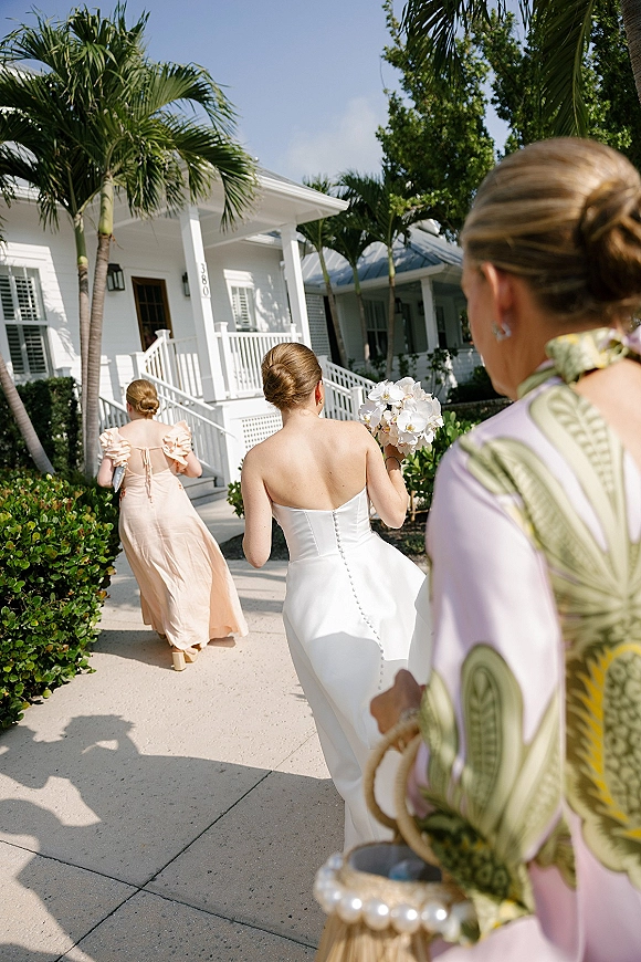 Bridal party arrival as bride in strapless satin gown holds white orchid bouquet, walking with bridesmaid by palm-lined white porch path