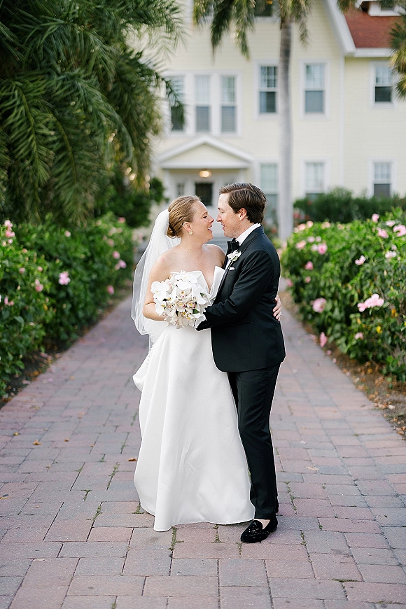 Couple portrait of bride and groom embrace, smiling on a brick garden walkway, bride in veil holding a white orchid bouquet