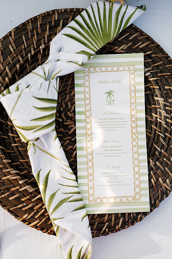 Wedding menu card with a palm leaf napkin on a wicker charger plate atop a white tablecloth, featuring green stripes and gold border