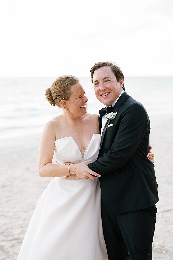 Couple portrait of bride and groom hugging at a beach wedding couple session, her strapless dress against his black tuxedo by the ocean horizon