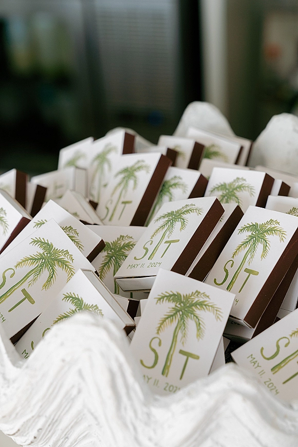 Wedding matchboxes with personalized matchboxes wedding favor sleeves, green typography and palm tree accents, stacked by a glass window and bowl