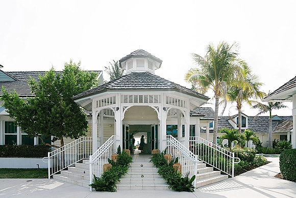Wedding venue entrance with greenery-lined stairs and lantern accents leading to a white gazebo pavilion framed by palm trees and sky