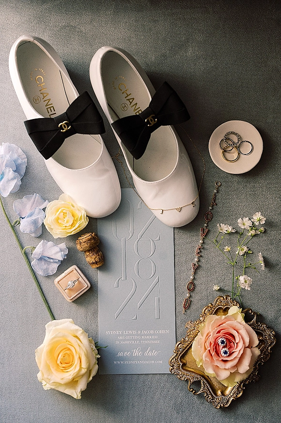 Wedding flat lay with wedding invitation flat lay, white bridal shoes with black bows, rings and jewelry arranged on gray fabric surface