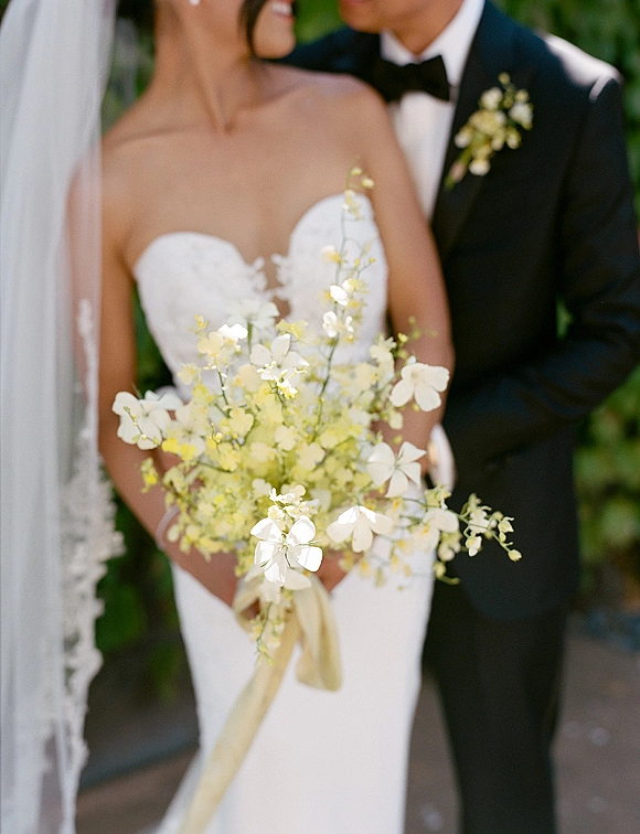 Wedding couple portrait of bride in strapless gown and veil holding a white and yellow bouquet beside groom in black tuxedo in garden foliage