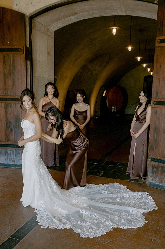 Bride getting ready as bridesmaids help bustle her strapless lace wedding dress, laughing in an arched hallway with pendant lights
