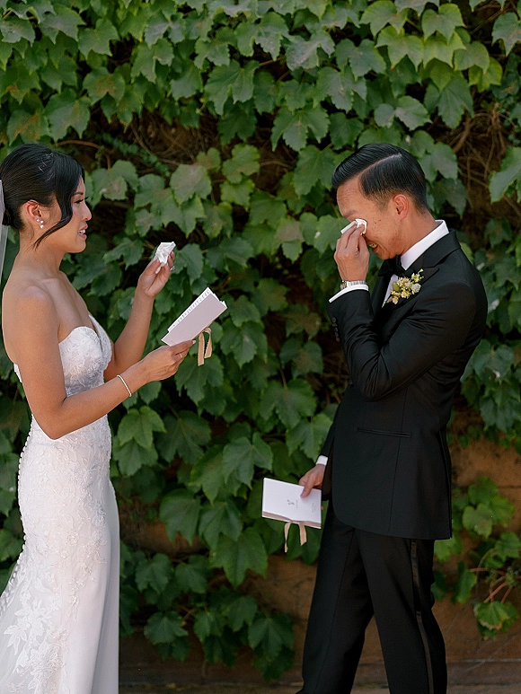 Wedding vows as bride reads from vow book to groom in tuxedo, veil trailing, ribbon bookmark visible against an ivy wall backdrop