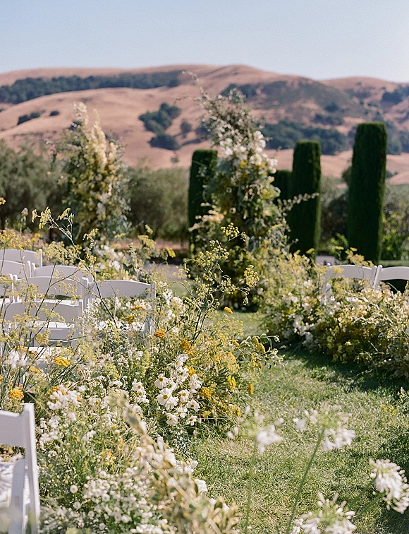 Ceremony aisle decor with wildflower aisle arrangements lining a grassy lawn, flanked by white folding chairs and cypress trees under blue sky