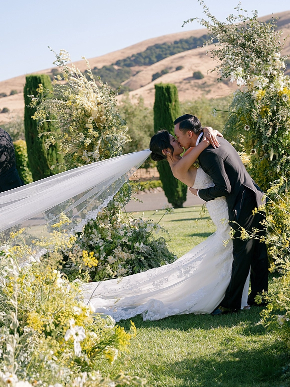 Wedding kiss portrait of bride and groom kissing under a floral arch, her long veil flowing over the lawn with cypress trees and hills behind