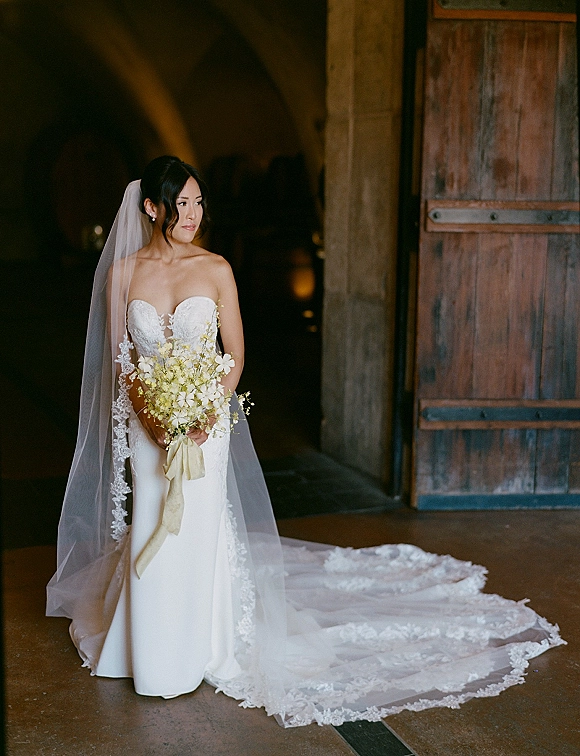 Bridal portrait of a bride in a strapless lace wedding dress holding a white bouquet before a wooden door and stone archway indoors
