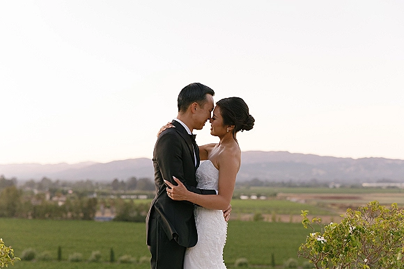 Couple portrait of newlyweds embracing with foreheads touching, bride in strapless dress and groom in tuxedo before a mountain valley backdrop