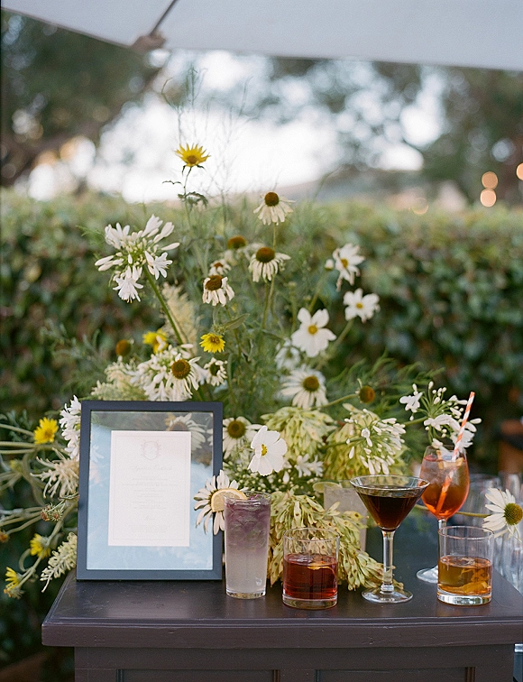 Cocktail table decor with a wildflower arrangement, framed menu sign, and cocktail glasses under bistro string lights by greenery hedges
