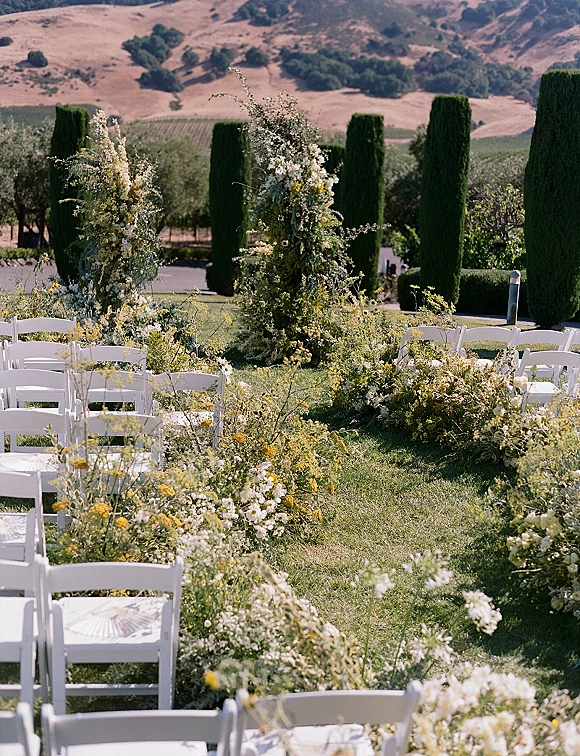 Ceremony aisle decor with meadow-style wildflower clusters and greenery lining white folding chairs on a vineyard lawn with cypress trees