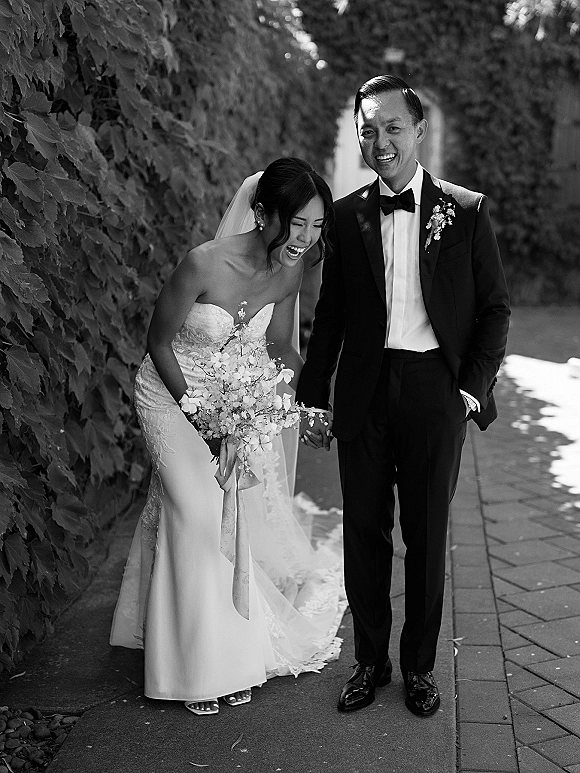 Couple portrait of bride and groom laughing, holding hands on an outdoor walkway by an ivy wall, her veil and bouquet ribbon flowing