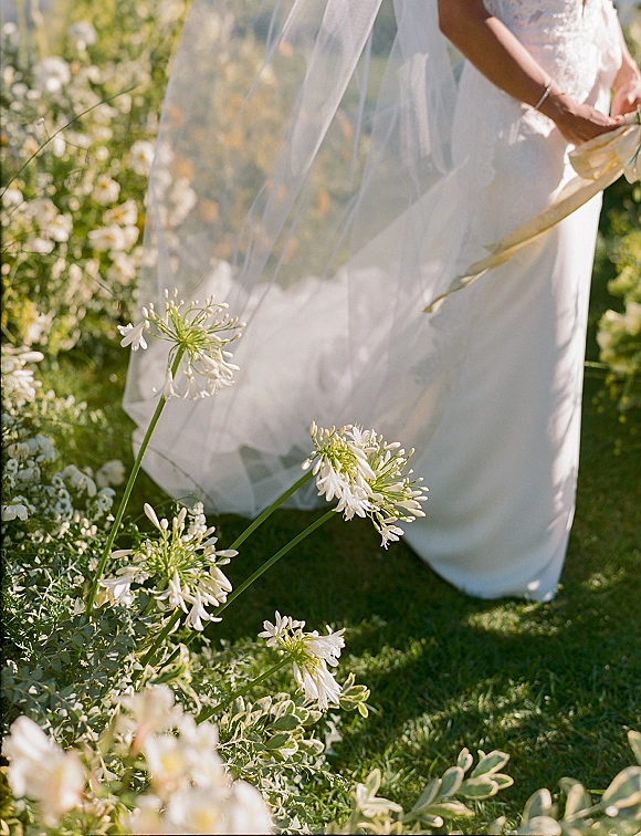 Wedding dress detail with a bridal veil draped over a simple gown, ribbon sash at the waist, and bouquet against garden foliage