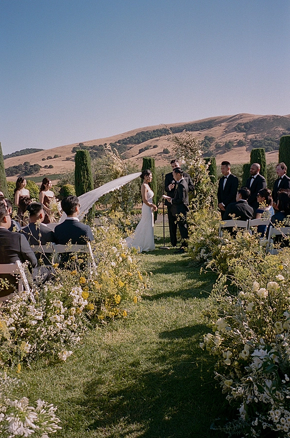 Wedding ceremony with couple exchanging vows under a draped white arch, floral-lined aisle on a garden lawn with hills and blue sky