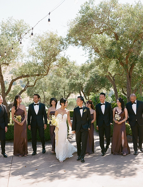 Wedding party portrait of bride and groom with wedding party walking on a sunlit paved path under trees, bouquets and string lights