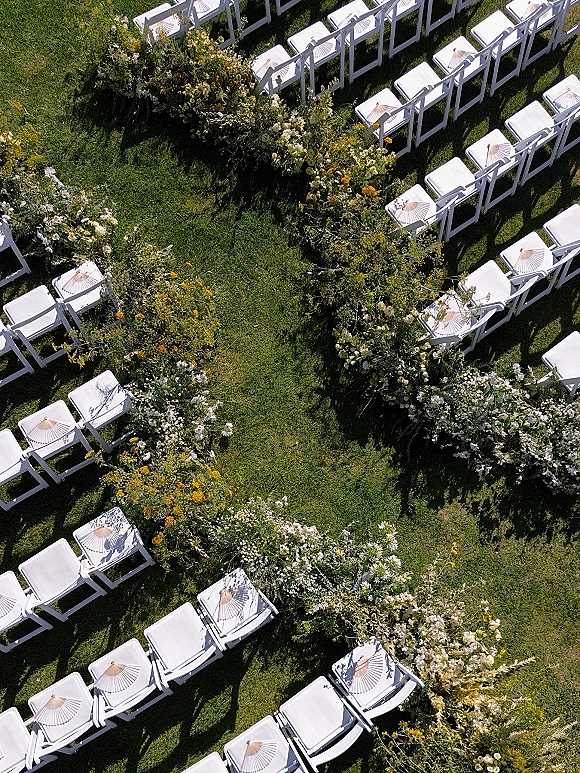 Ceremony aisle decor with floral lined aisle of yellow and white blooms beside white folding chairs and parasols on a grass lawn