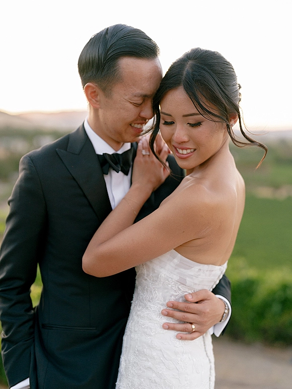 Couple portrait of bride and groom embrace with a forehead touch, her strapless lace dress and his black tuxedo in sunset hillside light