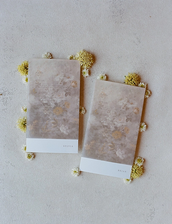 Wedding menu cards with floral print stationery and vellum overlays, tied with white belly bands and tiny yellow flowers on gray surface