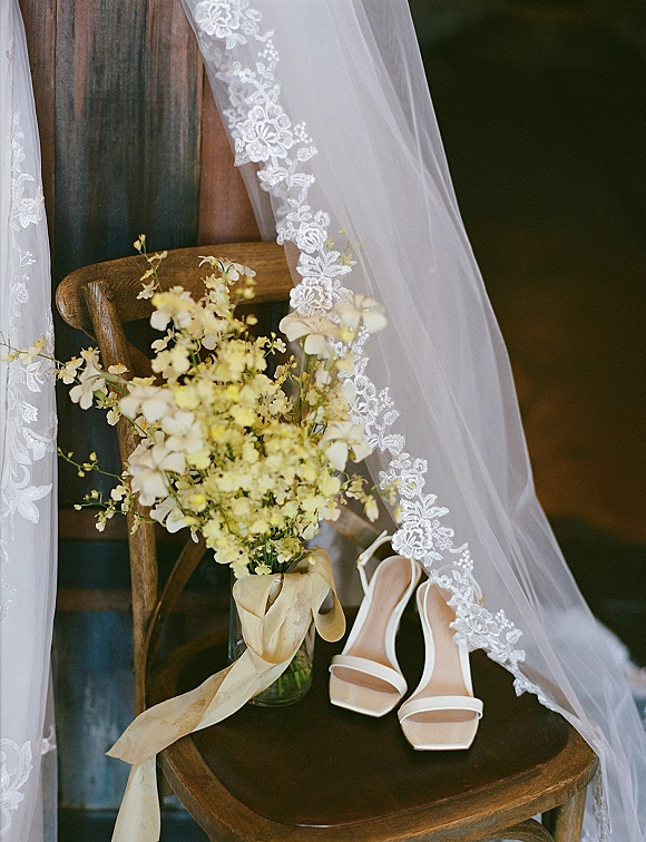 Bridal details with a lace veil and white strappy heels styled on a wooden chair beside a floral vase near a wood door