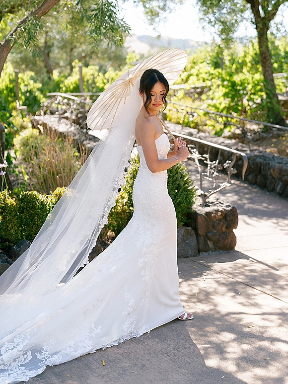Bridal portrait of a bride holding a white paper parasol in a strapless lace gown and long veil on a sunlit garden walkway by a stone wall