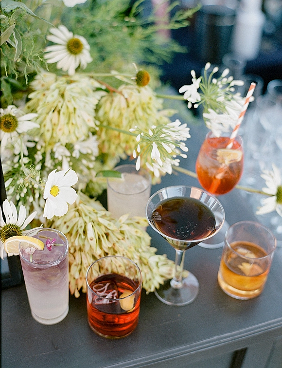 Wedding signature drinks in three cocktail glasses with ice, striped straw and lemon garnish beside daisy floral centerpiece on dark tabletop