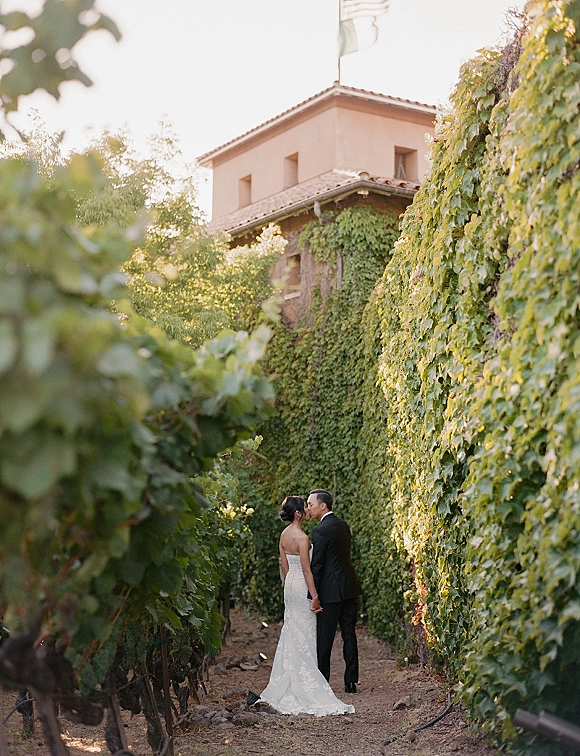 Wedding kiss portrait of bride in a strapless lace dress and groom in a tuxedo holding hands on a dirt path between vineyard rows