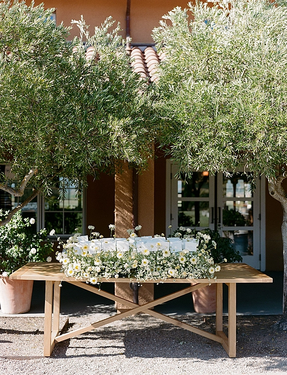 Wedding welcome table with white favor boxes and a daisy garland, set on a gravel patio by olive trees and a stucco building