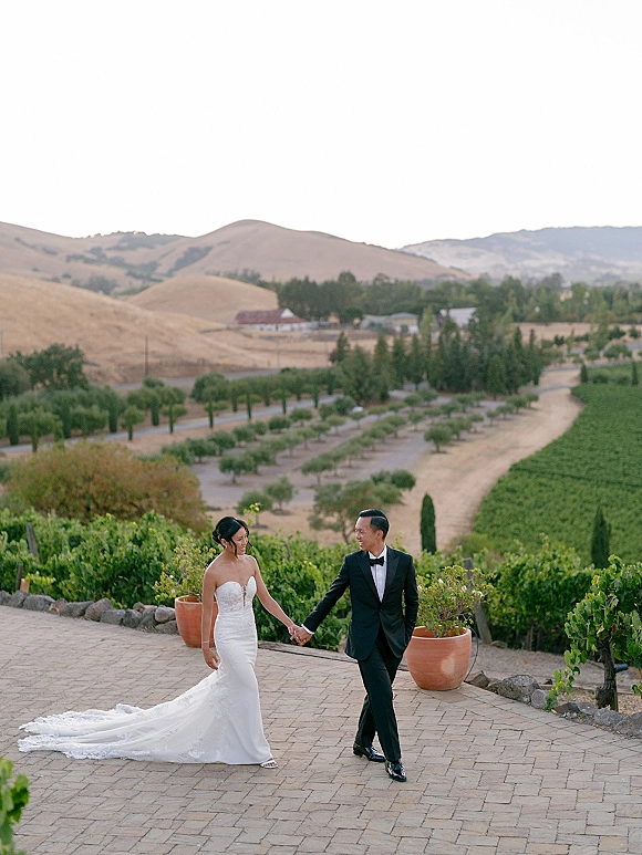 Couple portrait of bride and groom holding hands, walking on a courtyard with vineyard rows and hills behind, her long-train lace gown flowing