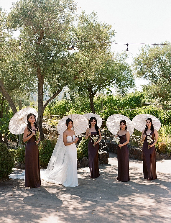 Bridal party portrait of the bride with bridesmaids under white parasols, bouquets in hand, on a stone path by olive trees and vineyard rows