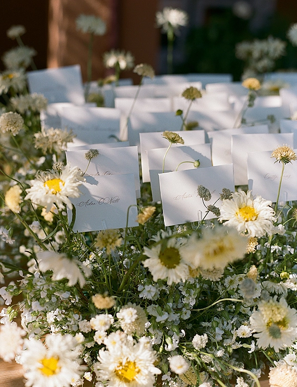 Wedding escort cards nestled among wildflowers in an escort card display, white calligraphy place cards on a sunlit wooden table in a garden setting
