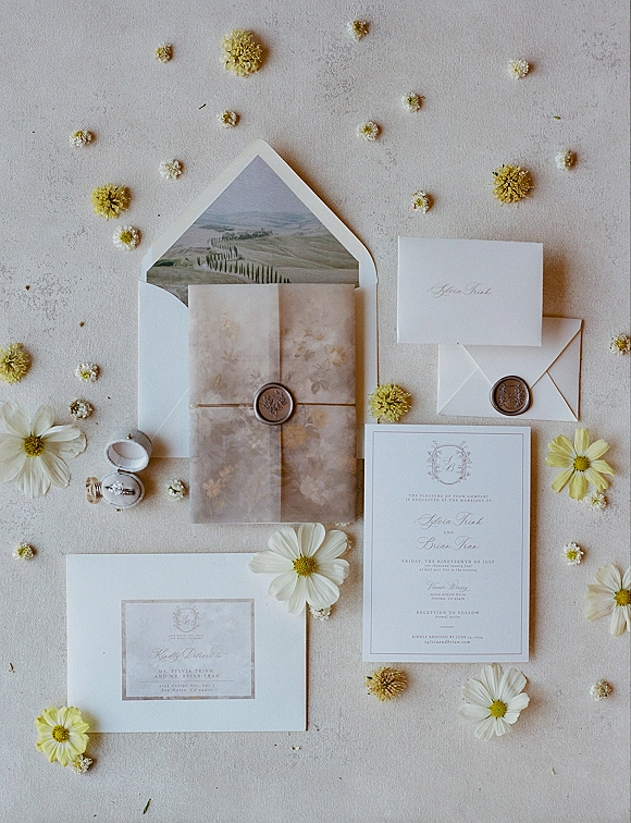 Wedding invitation suite with vellum wedding invitations, wax seals and twine, plus daisies and an engagement ring on a neutral surface