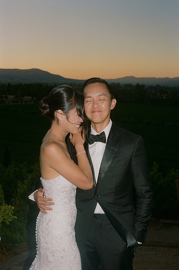 Couple portrait of bride and groom embracing, her strapless lace wedding dress and his tuxedo, against a sunset mountain garden backdrop