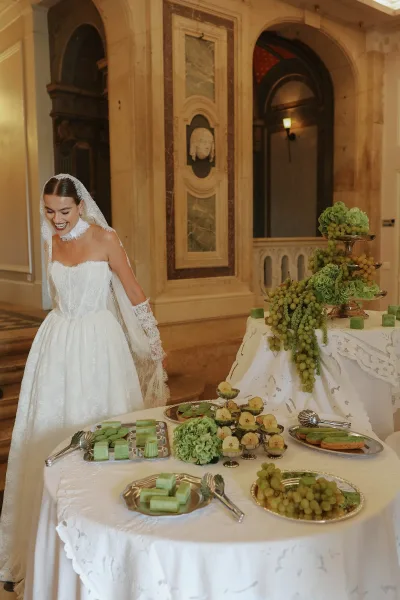 Bride portrait smiling in lace gloves and cathedral veil beside a wedding dessert table with green macarons, grapes, and candles in an ornate hall